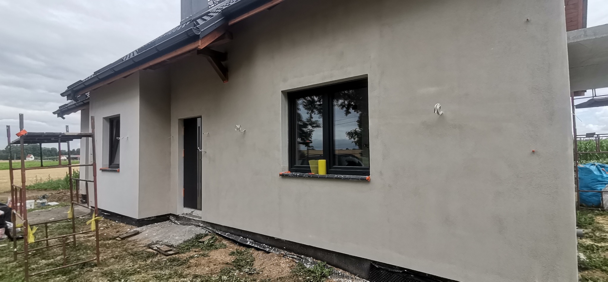 Świeżo otynkowana elewacja domu jednorodzinnego w odcieniach szarości, z widocznymi czarnymi ramami okiennymi i drzwiami, oraz żółtym kubkiem na parapecie. Na pierwszym planie fragment rusztowania...