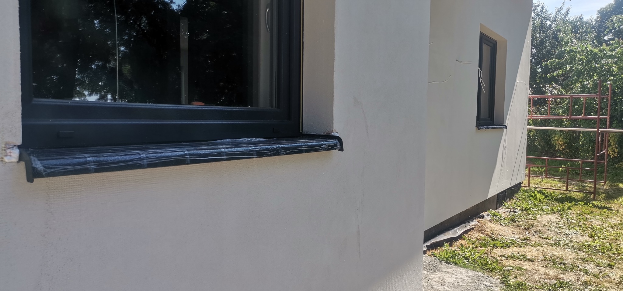 Fragment elewacji budynku z oknem z czarną ramą i parapetem zabezpieczonym folią ochronną, widoczne ślady po spływającej wodzie na świeżo pomalowanej ścianie i rusztowanie w tle.