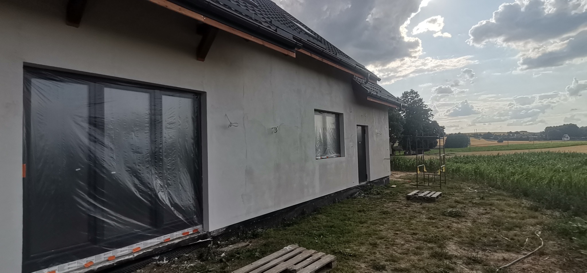 Surowa elewacja domu jednorodzinnego w trakcie prac, okna zabezpieczone folią, widoczne wyprowadzone kable i elementy mocujące na ścianie, w tle pola uprawne i pochmurne niebo.