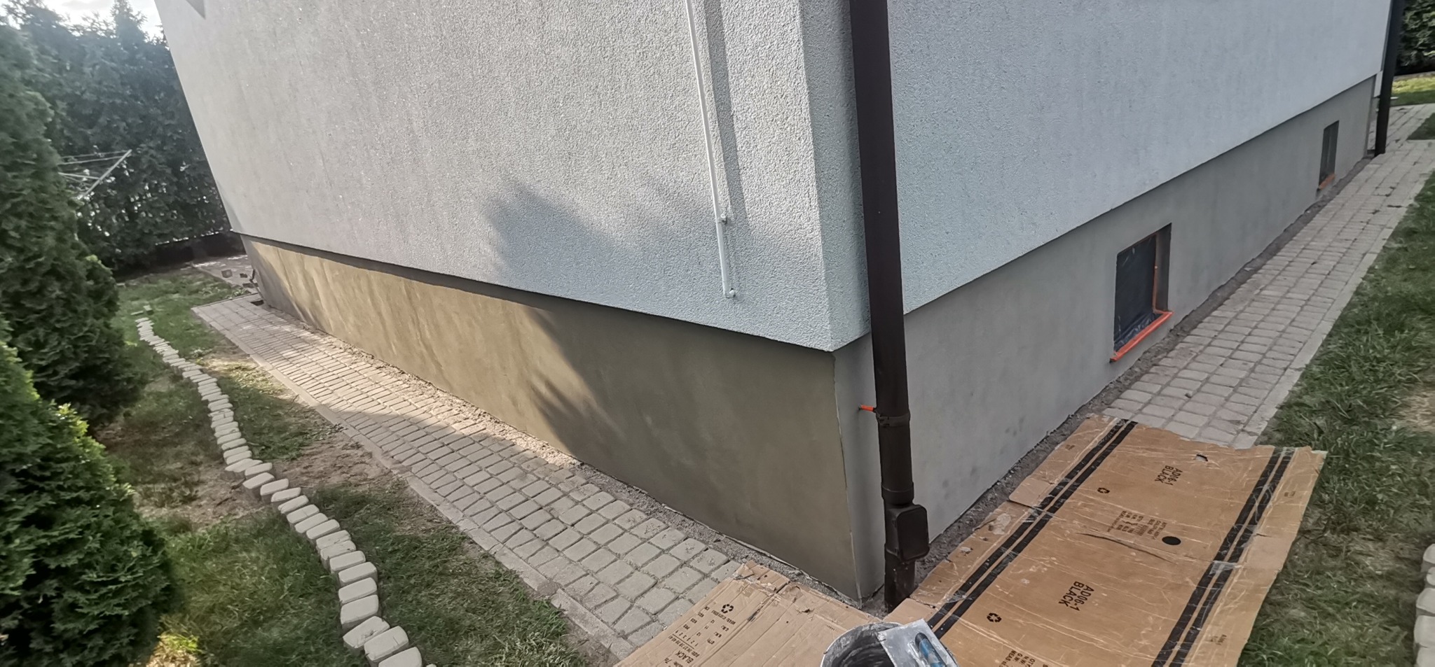 Świeżo pomalowana dolna część elewacji budynku w kolorze szarym, z widoczną rynną, zabezpieczonymi oknami i fragmentem chodnika z kostki brukowej.