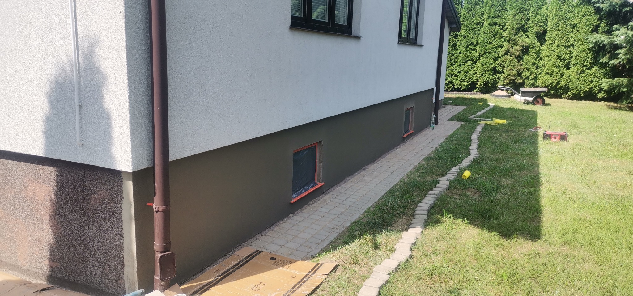 Fragment elewacji budynku po częściowym pomalowaniu, dolna część w kolorze grafitowym, okna zabezpieczone folią, na trawniku leżą żółte kubki i radio.