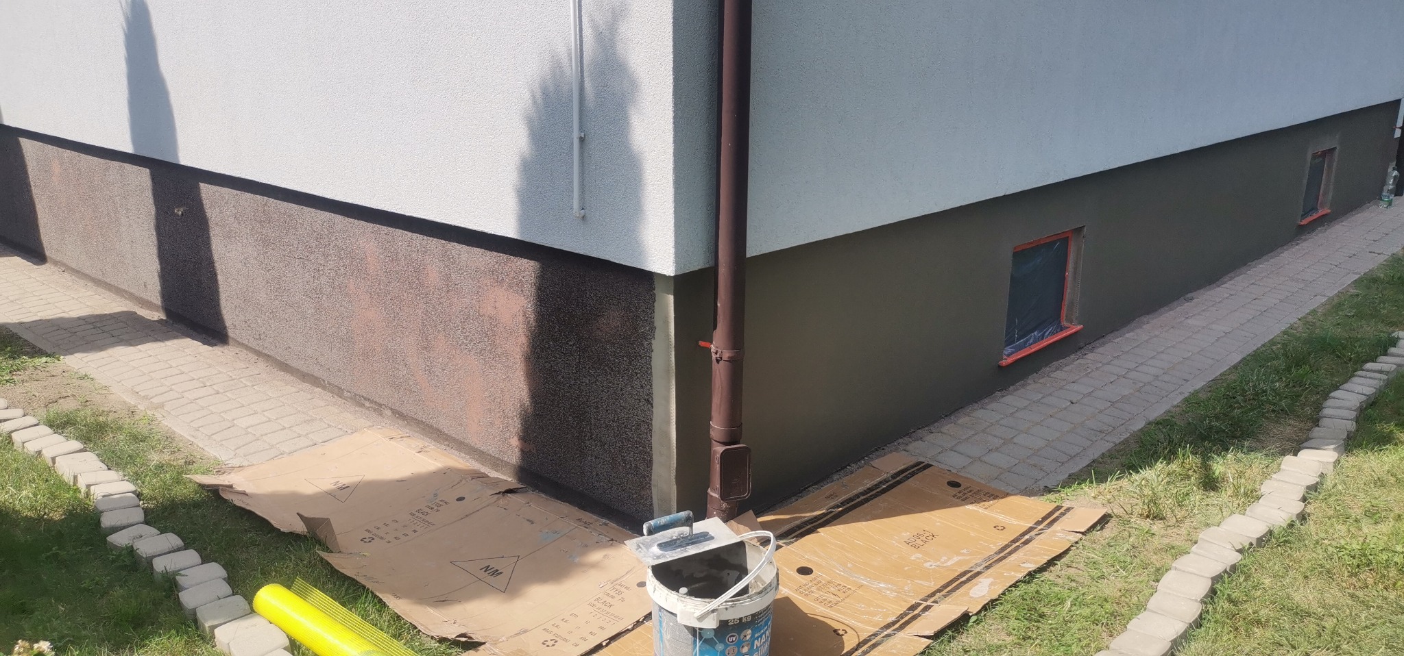 Fragment elewacji budynku z widocznym dwukolorowym wykończeniem, brązowym cokołem i jasną górną częścią. Okna zabezpieczone folią, na ziemi wiadro z zaprawą i narzędzia, zabezpieczone kartonem.
