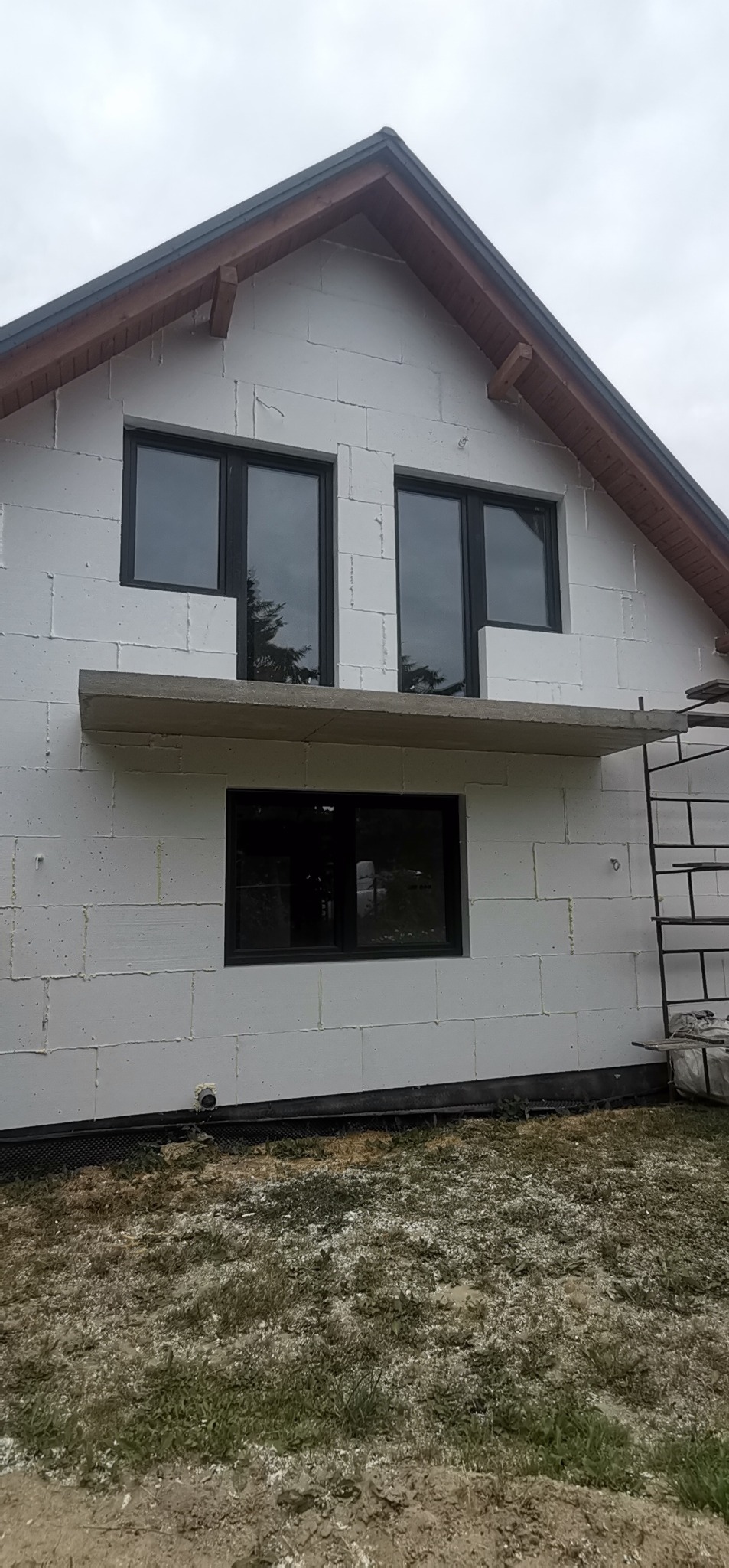 Niedokończona elewacja domu z białych bloczków, widoczne czarne ramy okienne i betonowy nadproże, obok stoi drabina.