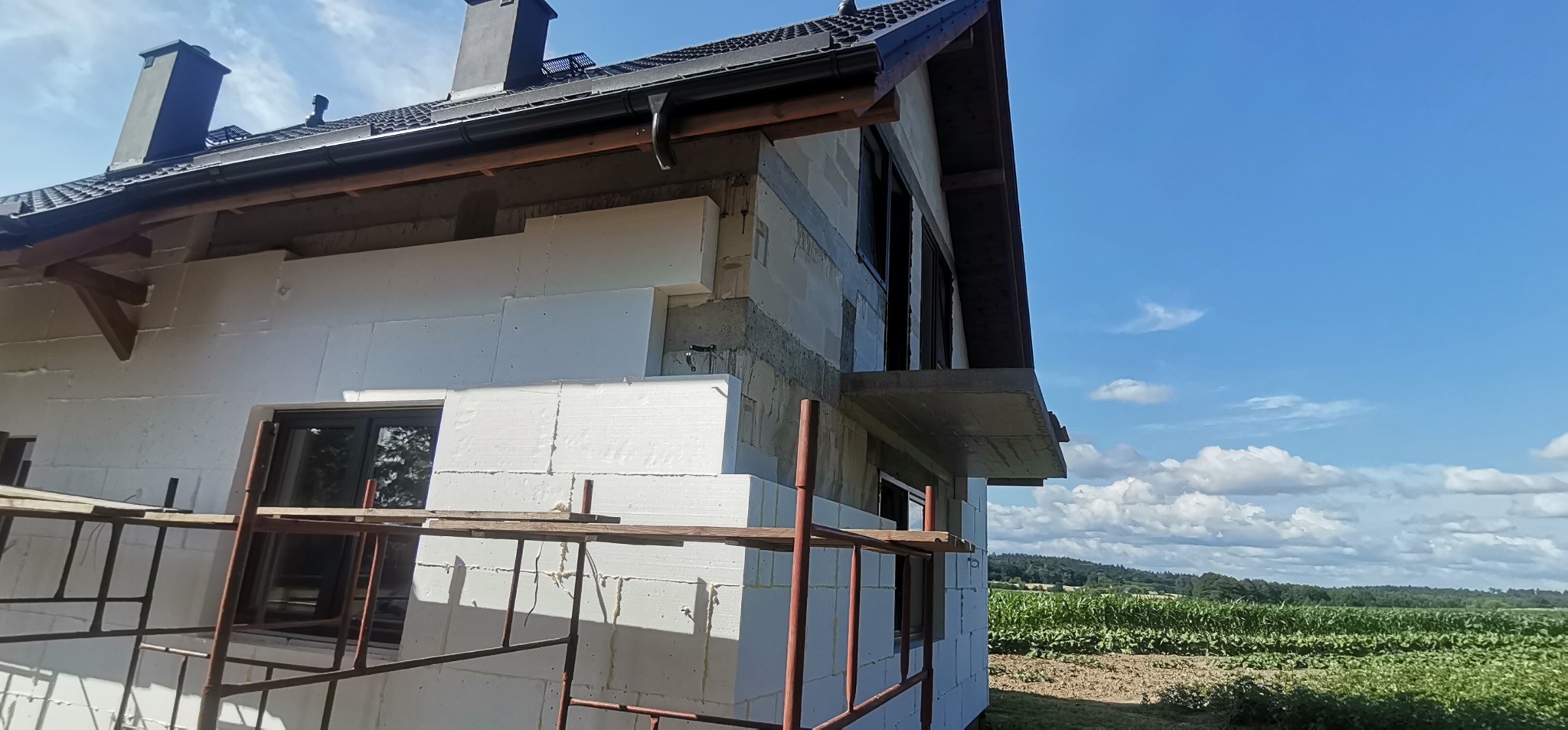 Dom w trakcie ocieplania styropianem, widoczne rusztowania, okna i fragment krajobrazu z polem w tle.