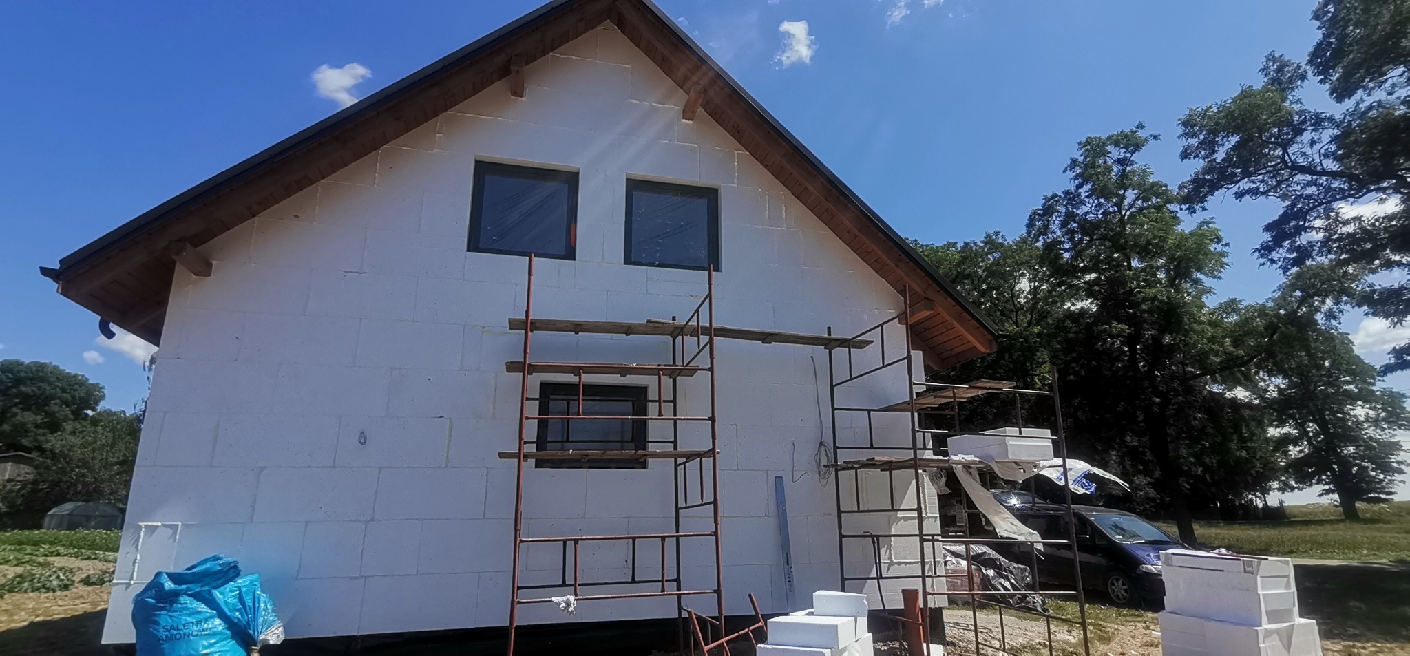 Dom w trakcie ocieplania styropianem, widoczne rusztowania, okna zafoliowane, worki z materiałami budowlanymi i samochód w tle.