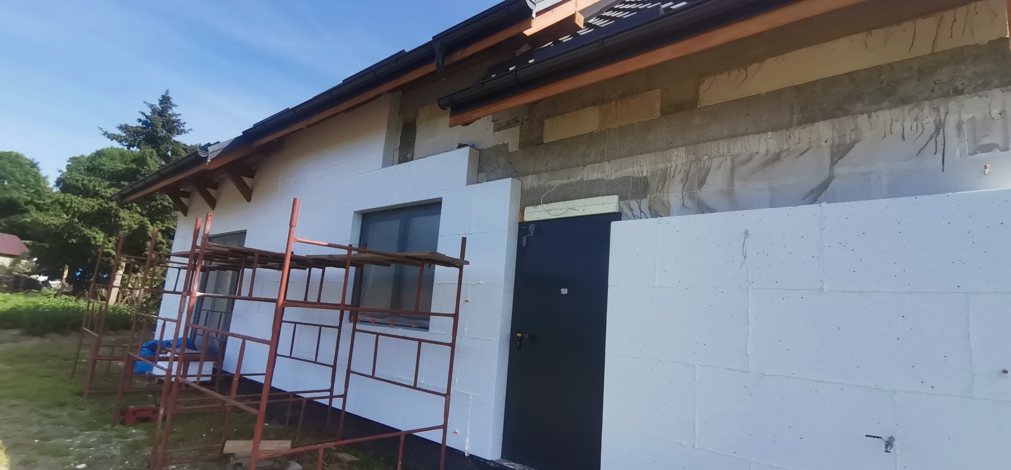Dom w trakcie ocieplania styropianem, widoczne rusztowanie, fragment niezabezpieczonej ściany i czarne drzwi wejściowe.