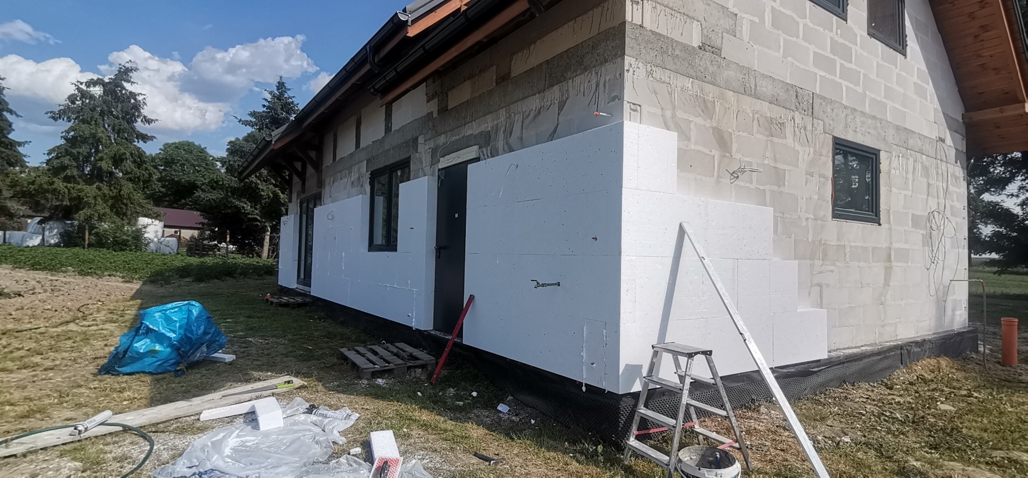 Dom w trakcie ocieplania styropianem, widoczna drabina, narzędzia i niebieski worek na śmieci na trawniku.