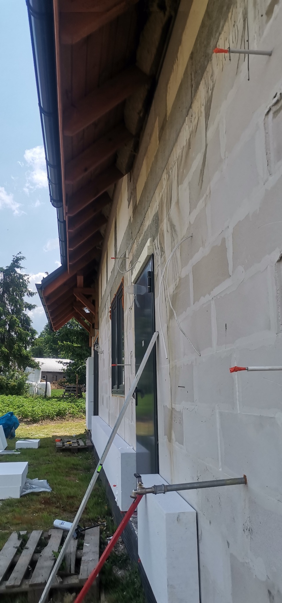 Ściana budynku w trakcie ocieplania styropianem, widoczne kołki mocujące, okna z zamontowaną stolarką, fragment dachu i rynny, trawa i zieleń w tle.