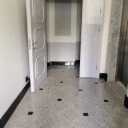 Korytarz wyłożony szarą, nakrapianą posadzką z czarnymi akcentami, prowadzący do otwartych drzwi i windy.