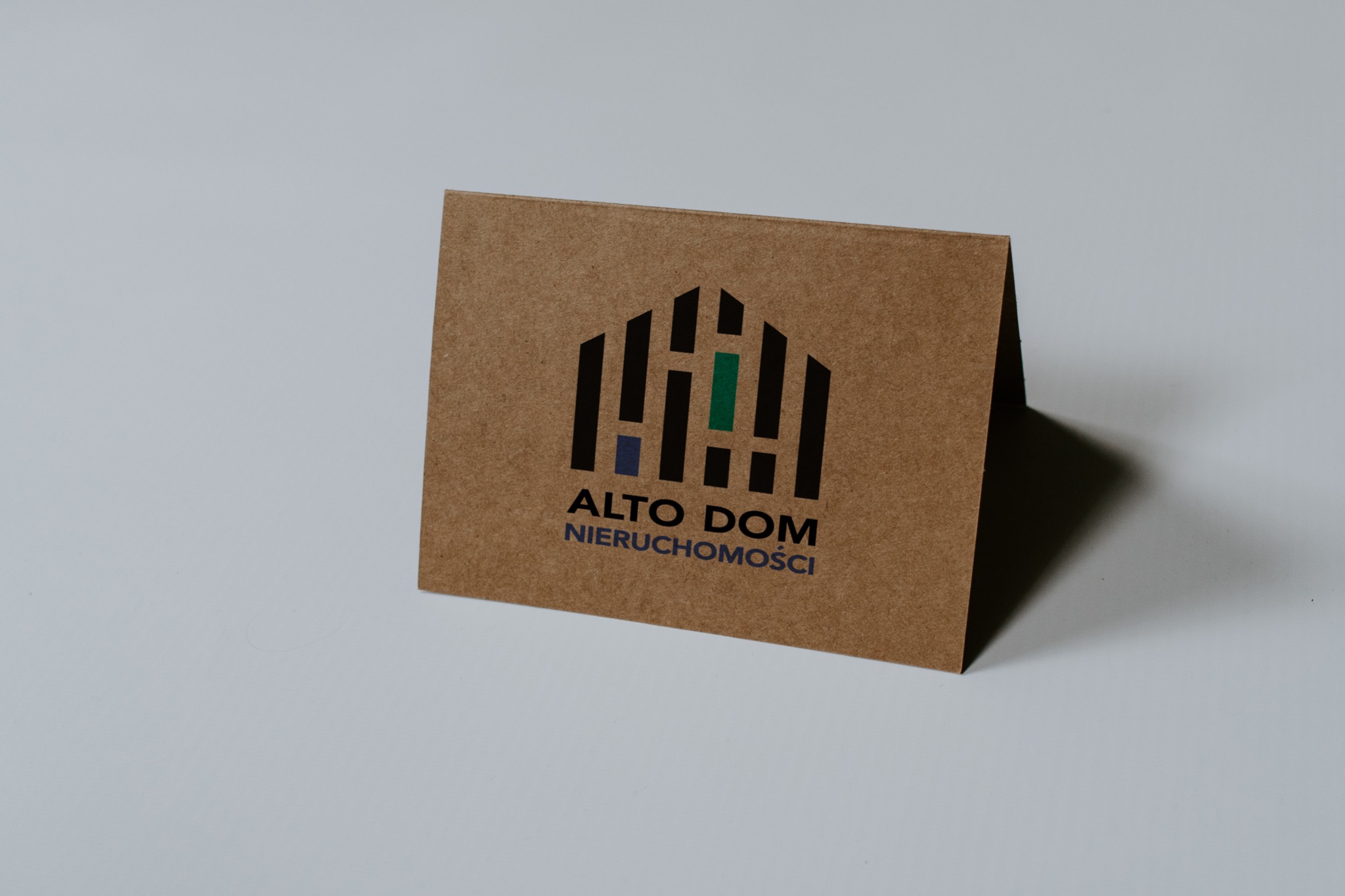 Brązowa, składana kartka z logo firmy 'Alto Dom Nieruchomości' w kolorach czarnym, niebieskim i zielonym na białym tle.