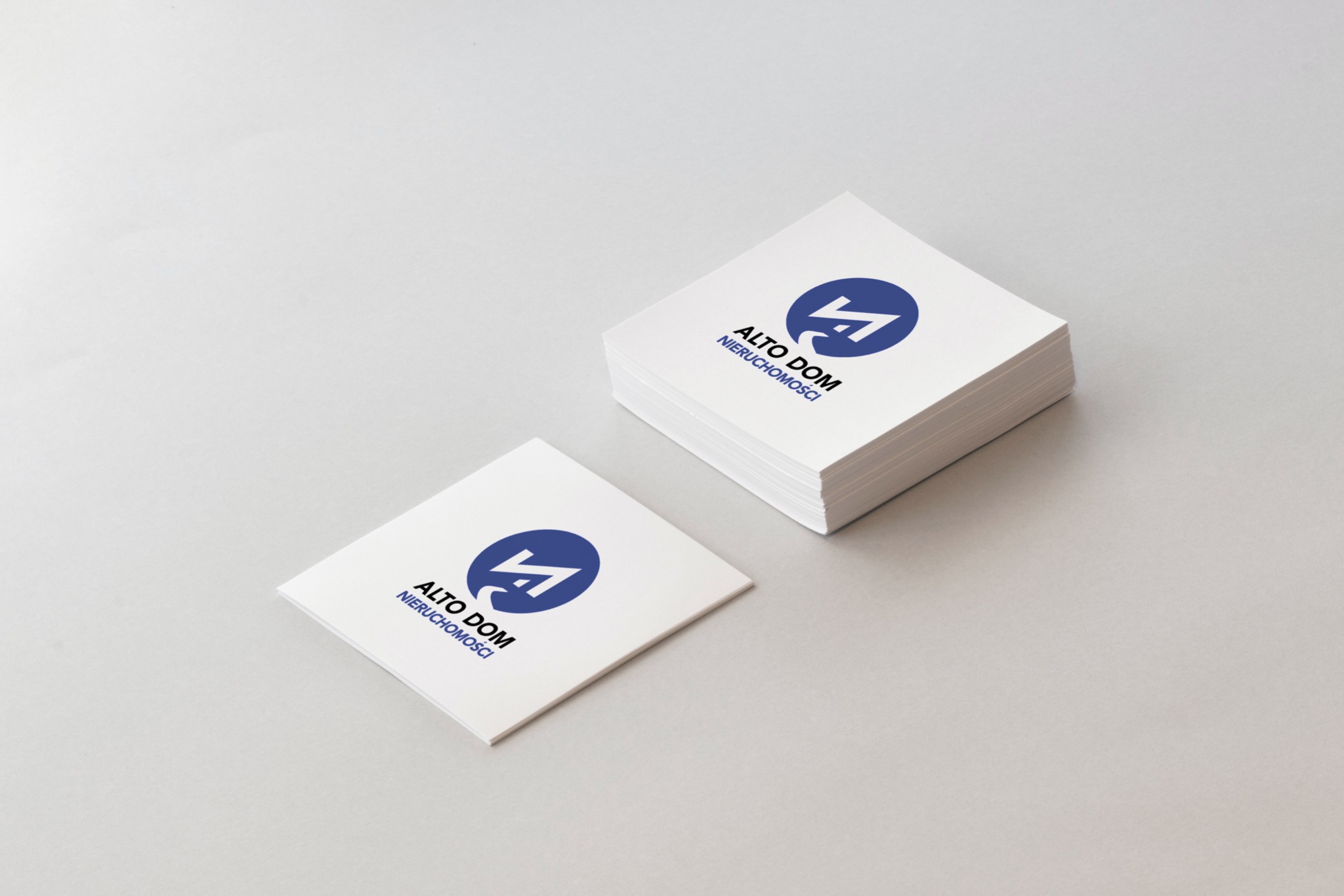 Stos wizytówek firmy Alto Dom Nieruchomości z niebieskim logo na białym tle, jedna wizytówka leży obok stosu.