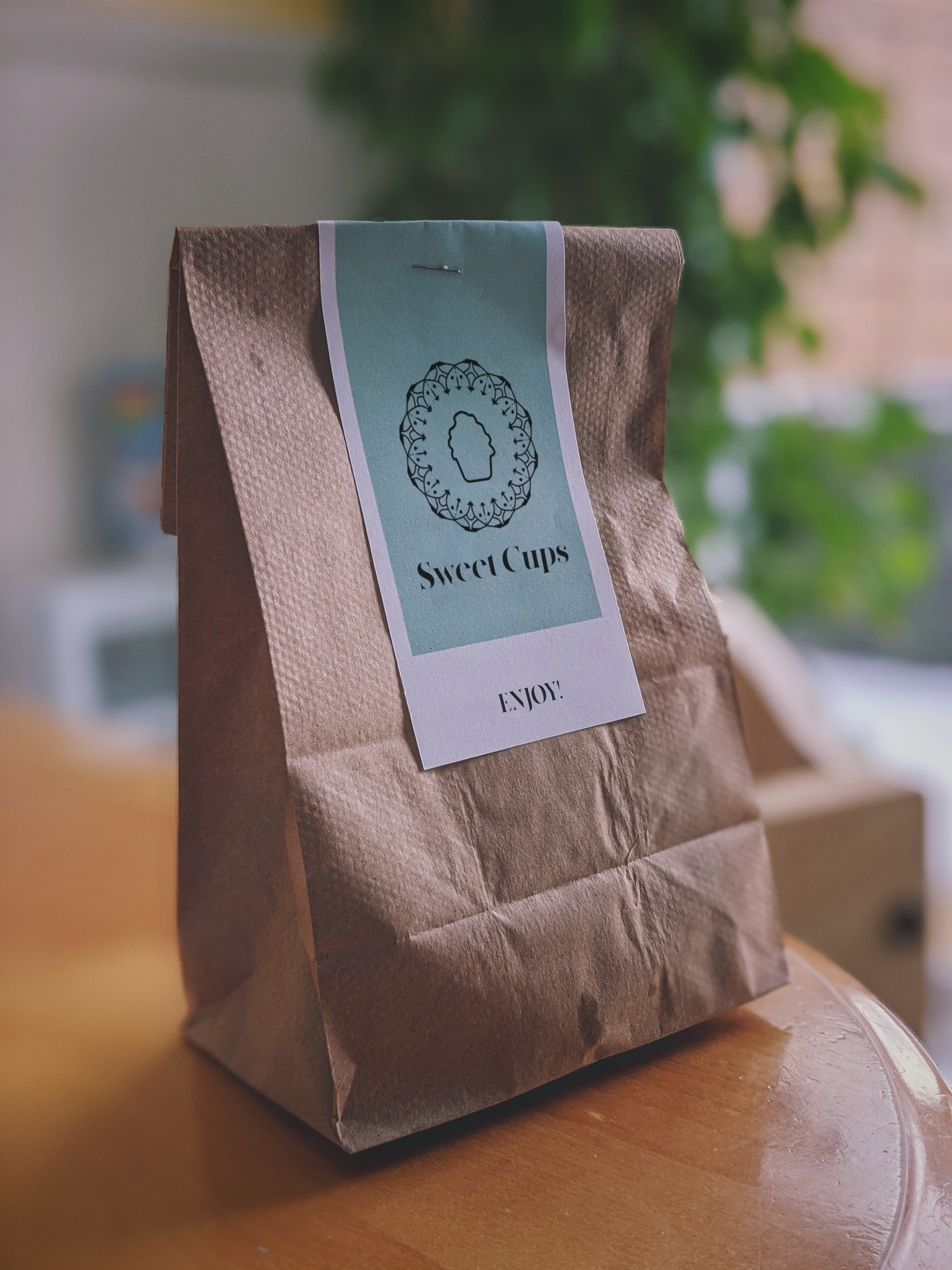 Brązowa papierowa torba z naklejoną etykietą z logo 'Sweet Cups' i napisem 'ENJOY!', przypięta zszywką, na drewnianym stole z rozmytym tłem roślinnym.