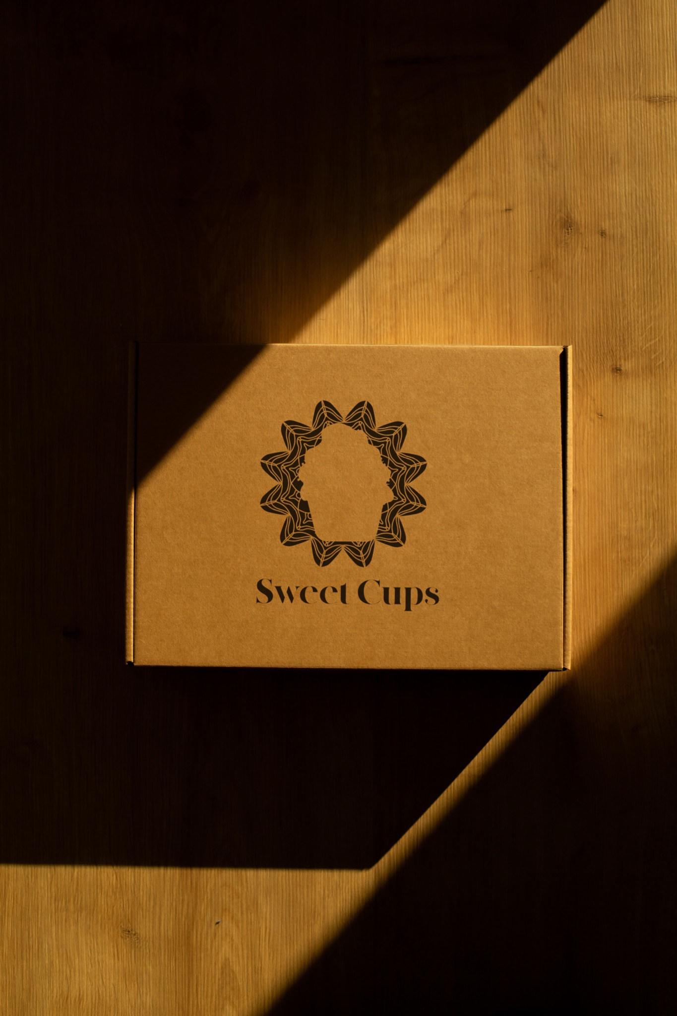Kartonowe pudełko z logo 'Sweet Cups' na drewnianym tle, oświetlone ukośnym promieniem słonecznym