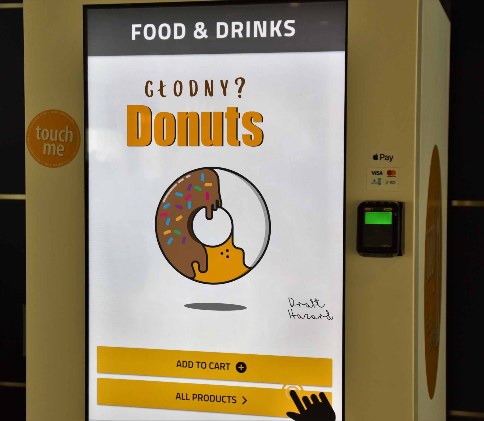 Automat sprzedający przekąski z ekranem dotykowym prezentującym graficzną ilustrację pączka z polewą i napisem 'Głodny? Donuts', opcje 'Dodaj do koszyka' i 'Wszystkie produkty', obok terminal...