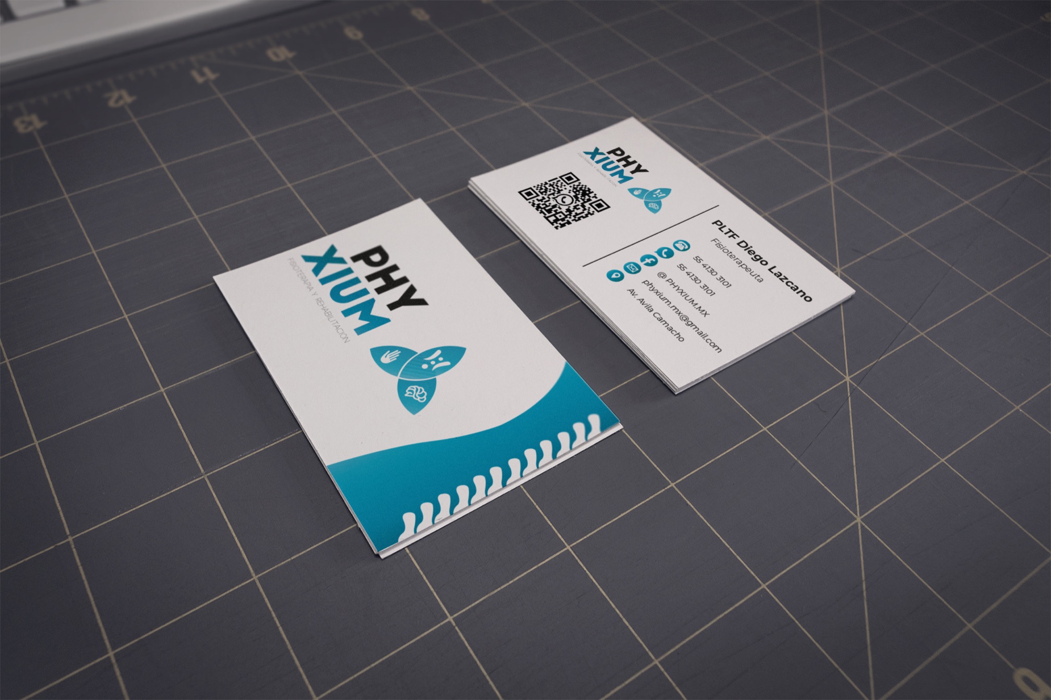 Dwie wizytówki firmy Phyxium, specjalizującej się w fizjoterapii i rehabilitacji, leżące na szarej macie do cięcia. Jedna wizytówka prezentuje logo i część graficzną, druga zawiera dane kontaktowe:...