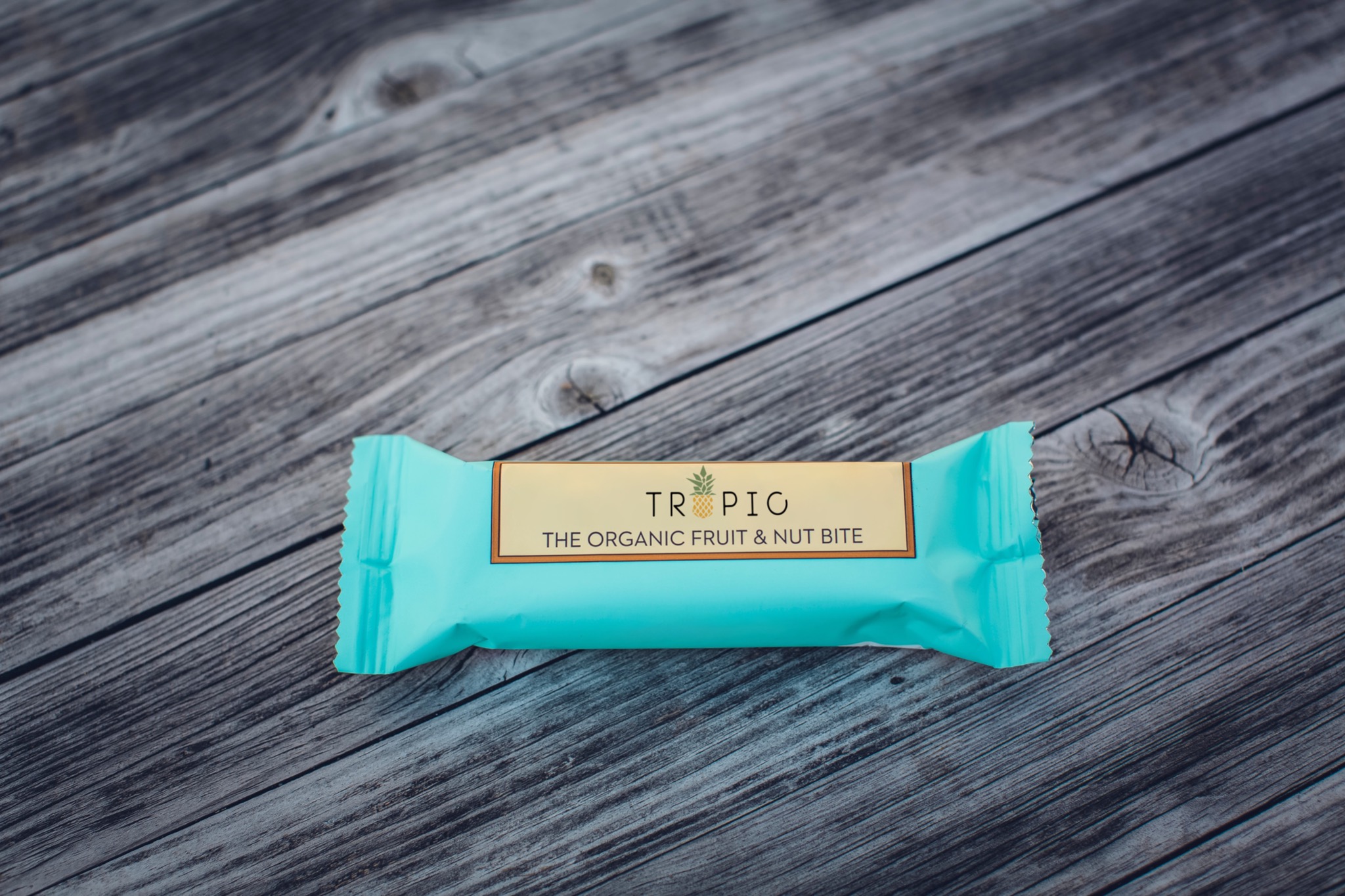 Turkusowe opakowanie batonika 'Tropic' z logo ananasa na szarym, drewnianym tle.