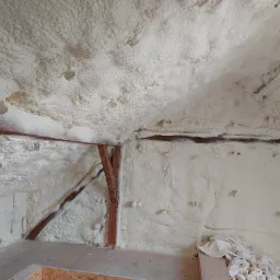 Poddasze z drewnianą konstrukcją pokryte grubą warstwą jasnej pianki izolacyjnej, widoczne nierówności i struktura pianki, fragment podłogi z płyty OSB.