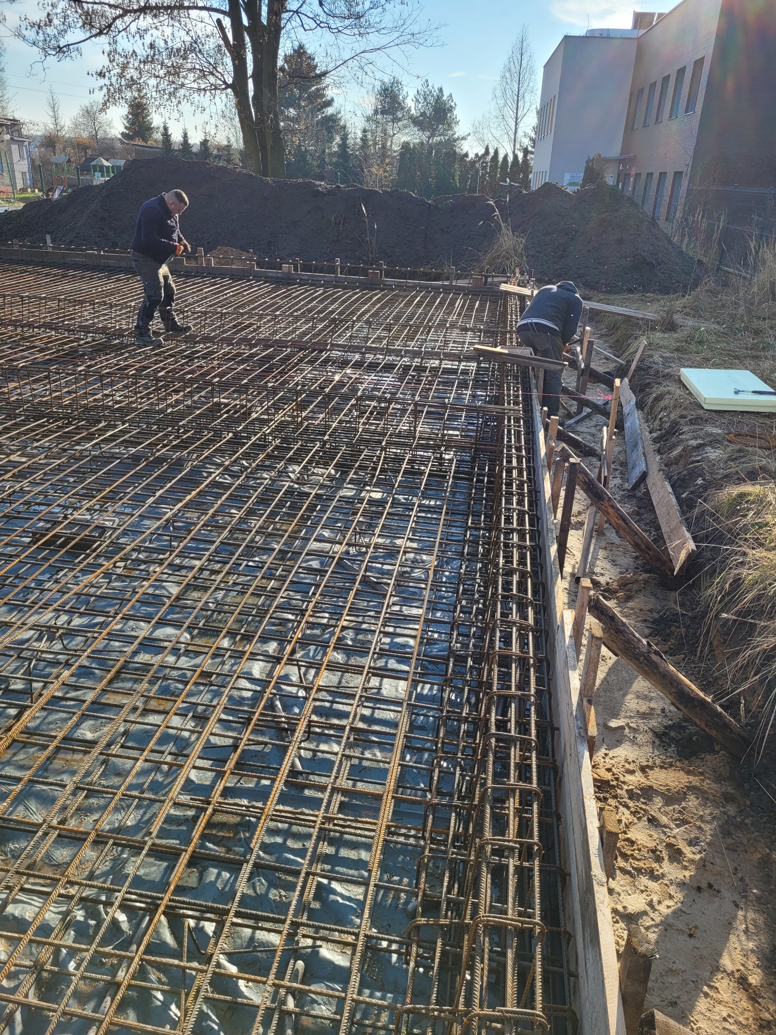 Przygotowanie zbrojenia fundamentów pod budynek, widoczni dwaj pracownicy w trakcie pracy, drewniane szalunki i wykop pod fundamenty.