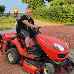 Kameleon Usługi Sprzątające i Ogrodnicze - Mężczyzna siedzi na czerwonej kosiarce traktorku Kubota GR1600II na tle zieleni i budynku, z włączonym pomarańczowym światłem ostrzegawczym.