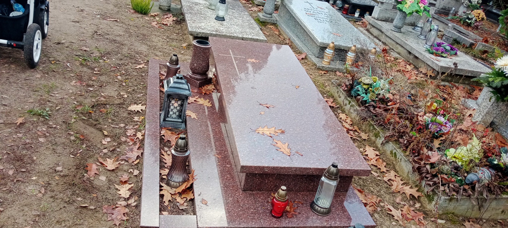 Czerwony granitowy nagrobek z lampionami i zwiędłymi liśćmi dębu na cmentarzu, widok z góry.