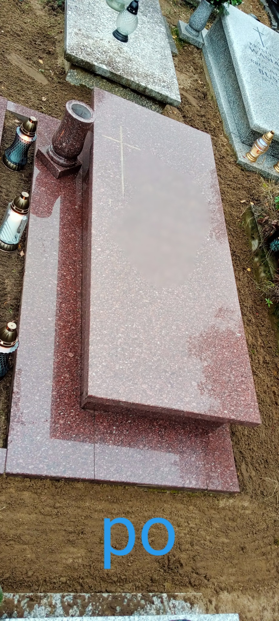 Nowy, bordowo-szary nagrobek z granitu z krzyżem, wazonem i lampionami, otoczony świeżo ubitą ziemią.