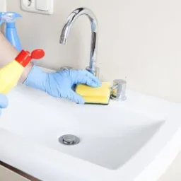 Osoba w niebieskich rękawiczkach czyści białą umywalkę żółtą gąbką i płynem w żółtej butelce z czerwonym dozownikiem, widoczny także niebieski spray.