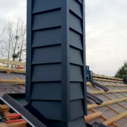 Czarny, metalowy komin wentylacyjny na dachu w trakcie montażu, widoczne elementy konstrukcji dachu i narzędzia dekarskie.