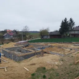 Fundamenty w trakcie budowy, widok z góry na teren budowy z częściowo wzniesionymi ścianami fundamentowymi z bloczków betonowych, drewnianym szalunkiem, z pracownikiem w niebieskiej kurtce w tle...