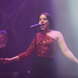 MARCIN STRUS Agencja Artystyczna M-art - Wokalistka w cekinowej bluzce śpiewa do mikrofonu na scenie, w tle klawiszowiec i fioletowe światła