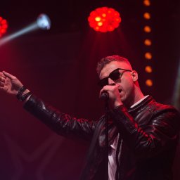 MARCIN STRUS Agencja Artystyczna M-art - Wokalista w czarnej skórzanej kurtce i okularach przeciwsłonecznych śpiewa do mikrofonu na scenie oświetlonej czerwonymi i pomarańczowymi światłami.