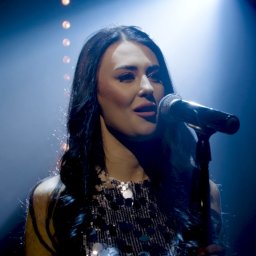 MARCIN STRUS Agencja Artystyczna M-art - Wokalistka z długimi ciemnymi włosami śpiewa do mikrofonu na scenie w niebieskim oświetleniu punktowym, ubrana w błyszczącą sukienkę.