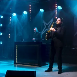 Wokalista grający na saksofonie na scenie z DJ-em w tle, oświetlenie sceniczne w kolorze niebieskim i białym, logo zespołu w prawym górnym rogu.