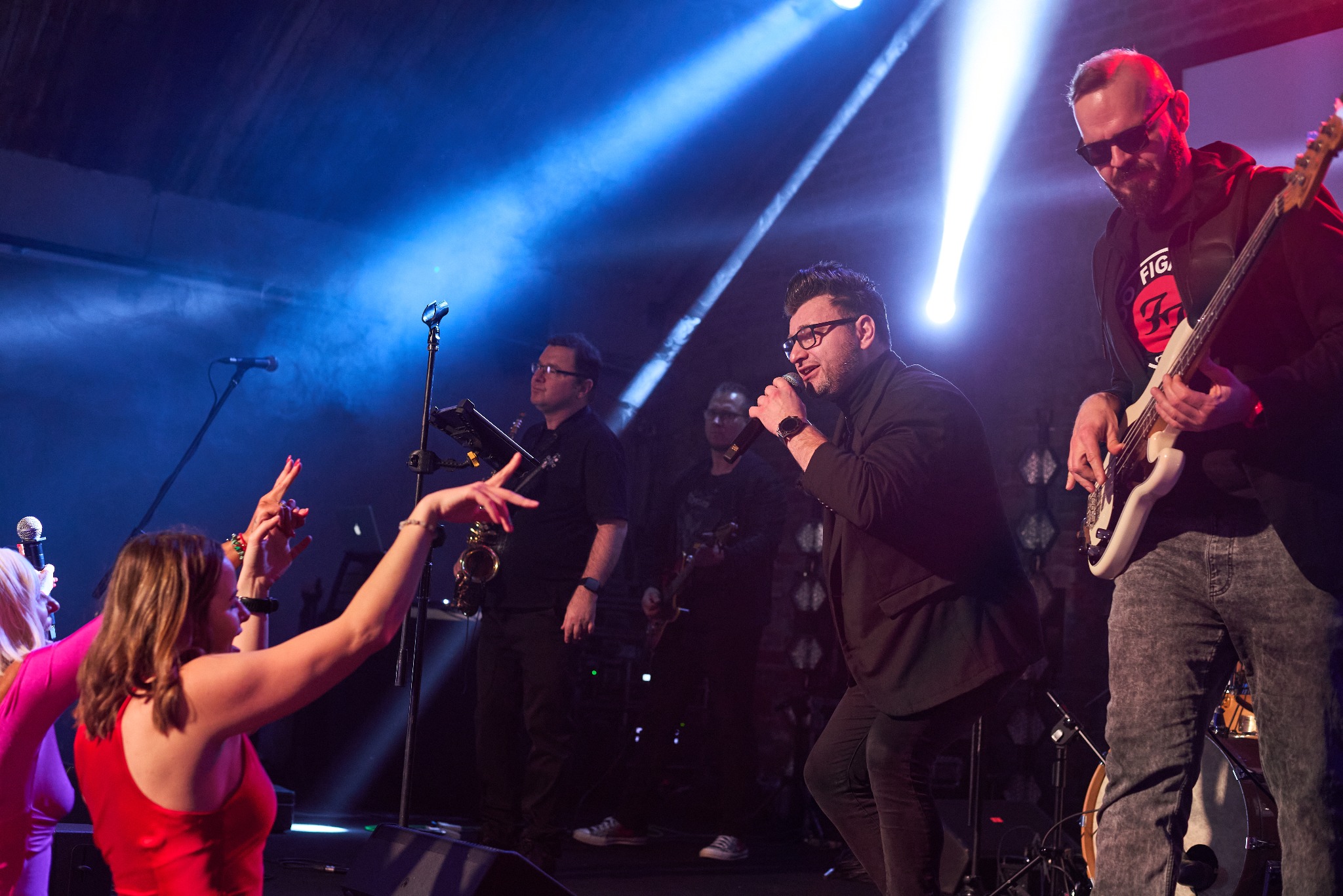 Cover band M-ART LIVE na scenie: wokalista w czarnym garniturze śpiewa do mikrofonu, obok saksofonista i basista w okularach przeciwsłonecznych grają, a publiczność entuzjastycznie reaguje.