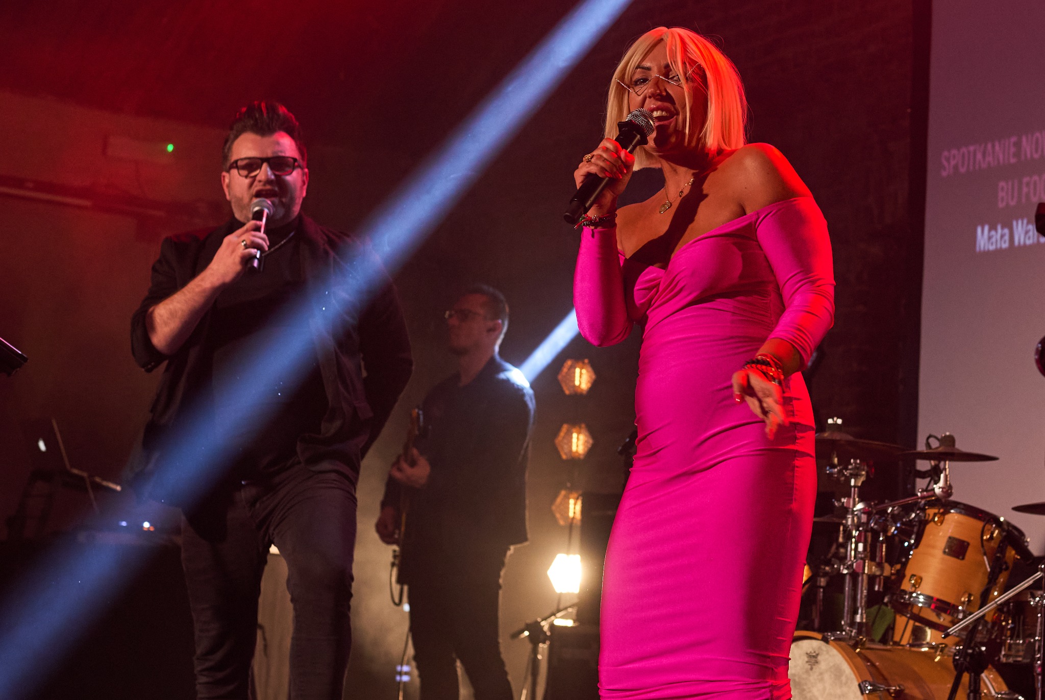 Wokalistka w różowej sukience śpiewa na scenie z mikrofonem, obok mężczyzna w okularach również śpiewa, w tle widoczny gitarzysta i perkusja, oświetlenie sceniczne.