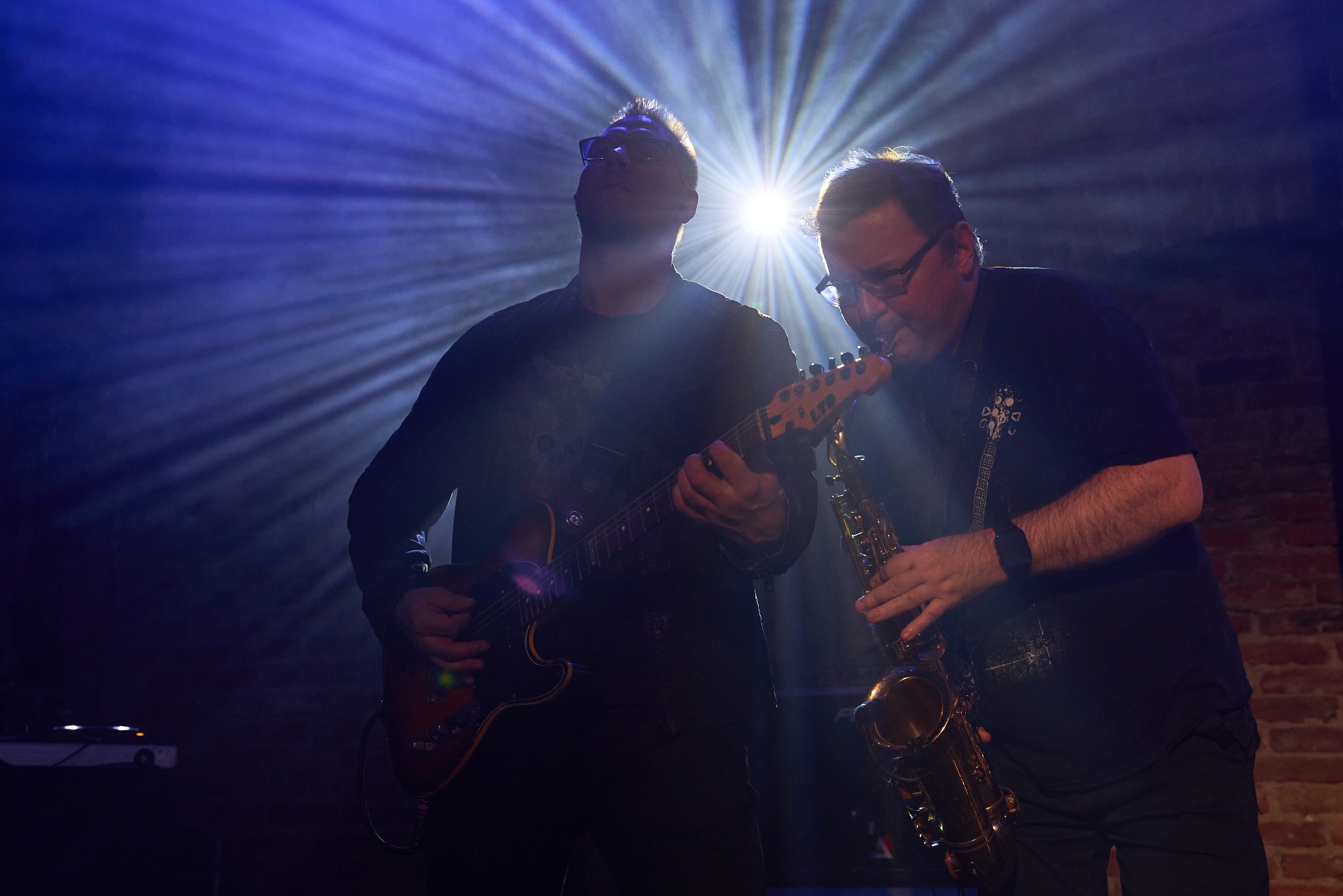 Dwóch muzyków w trakcie występu na scenie oświetlonej niebieskim światłem punktowym. Jeden gra na gitarze elektrycznej, drugi na saksofonie.