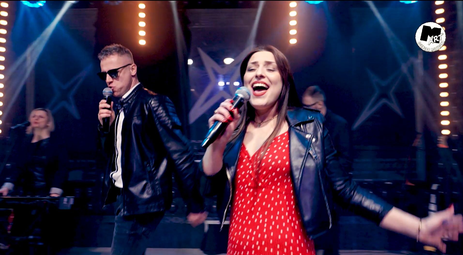 Występujący na scenie wokalista w okularach i wokalistka w czerwonej sukience w białe kropki, oboje w skórzanych kurtkach, śpiewają do mikrofonów na tle sceny z oświetleniem.