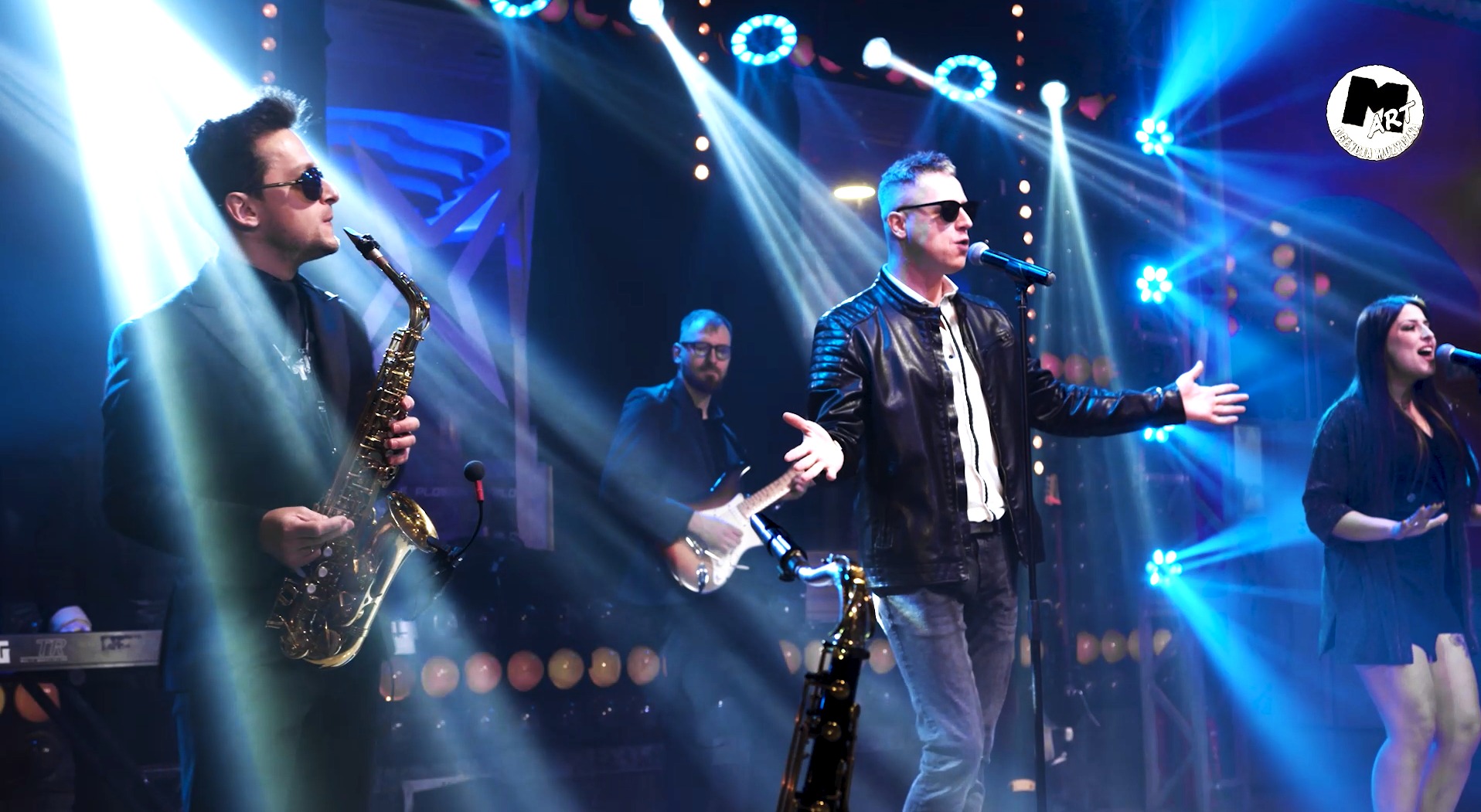 Występujący na scenie zespół: saksofonista w ciemnych okularach i garniturze, wokalista w skórzanej kurtce, gitarzysta z brodą oraz wokalistka, oświetleni niebieskimi reflektorami.