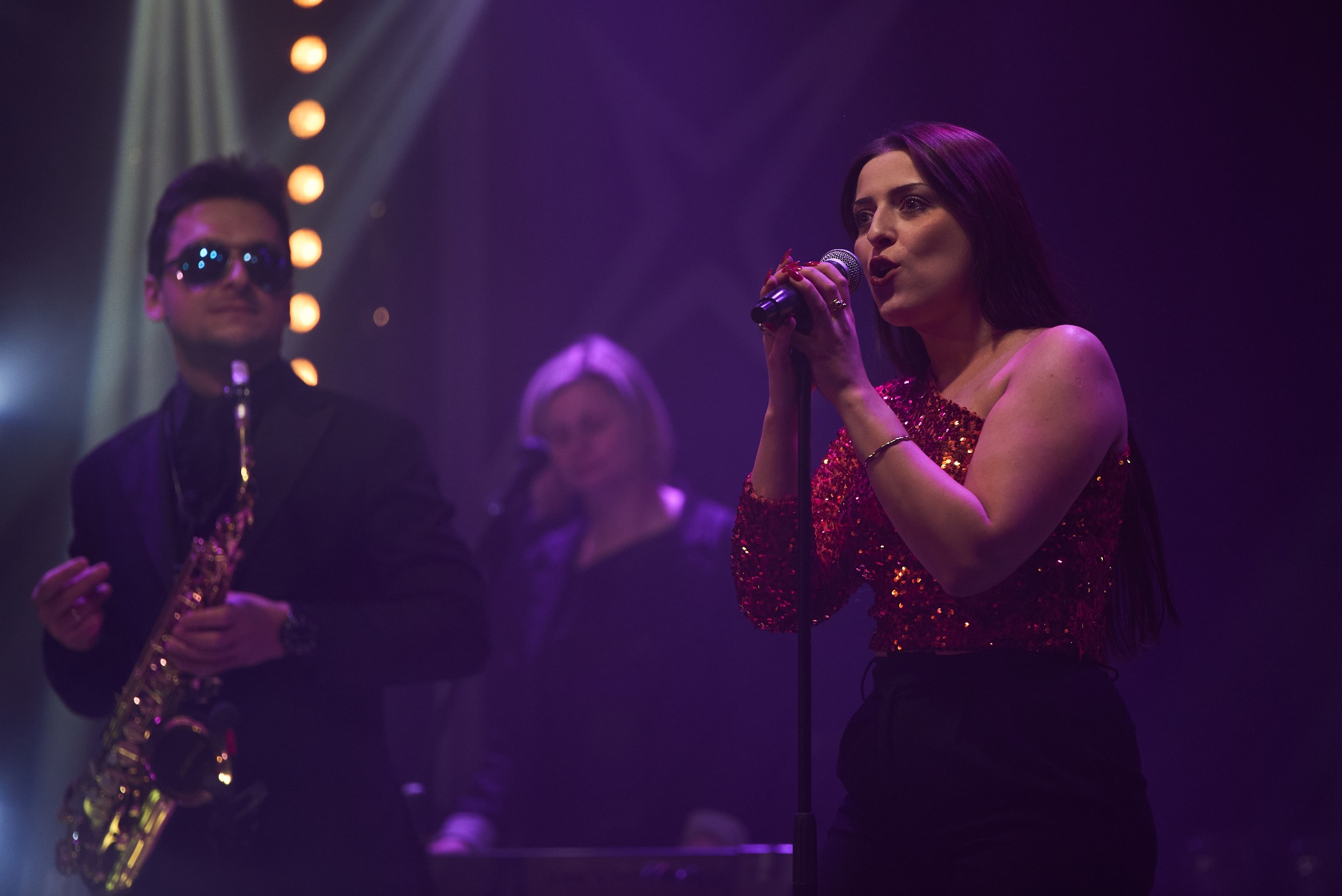 Wokalistka w cekinowej bluzce śpiewa do mikrofonu, obok saksofonista w okularach przeciwsłonecznych, w tle klawiszowiec, scena oświetlona fioletowymi światłami.