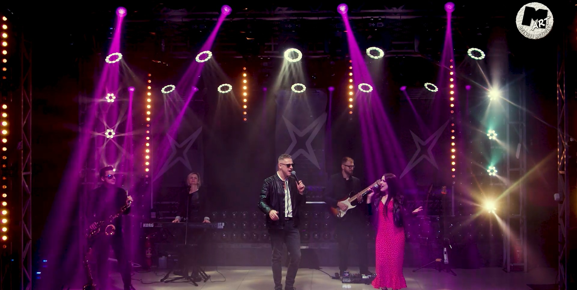 Występ na scenie: wokalista w skórzanej kurtce, saksofonista, gitarzysta i wokalistka w czerwonej sukience, podświetleni fioletowymi reflektorami.