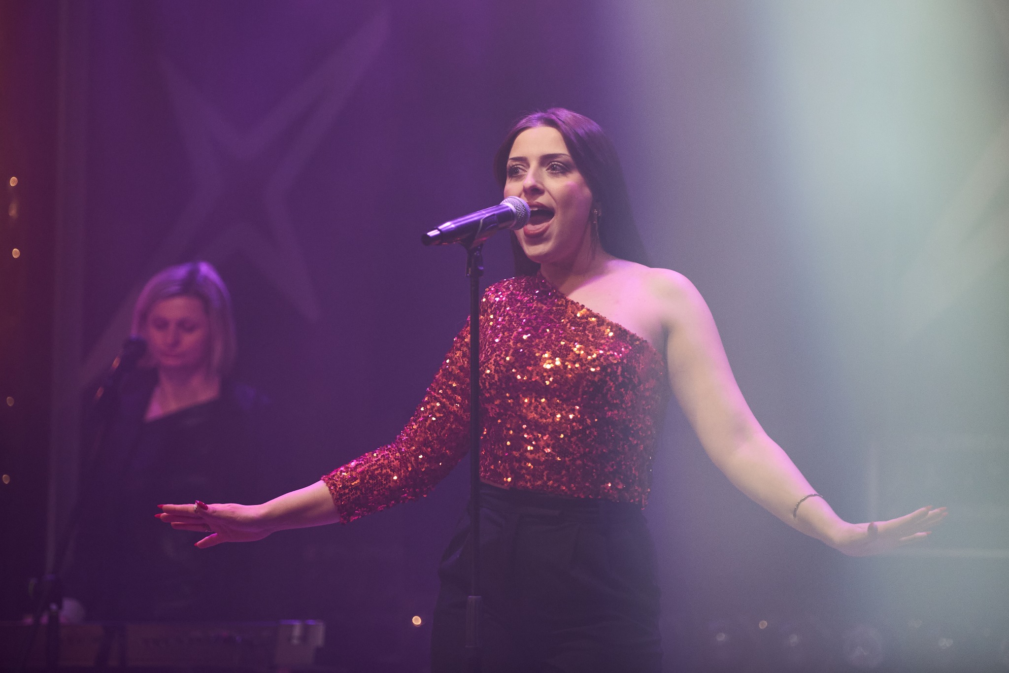 Wokalistka w cekinowej bluzce śpiewa do mikrofonu na scenie, w tle klawiszowiec i fioletowe światła