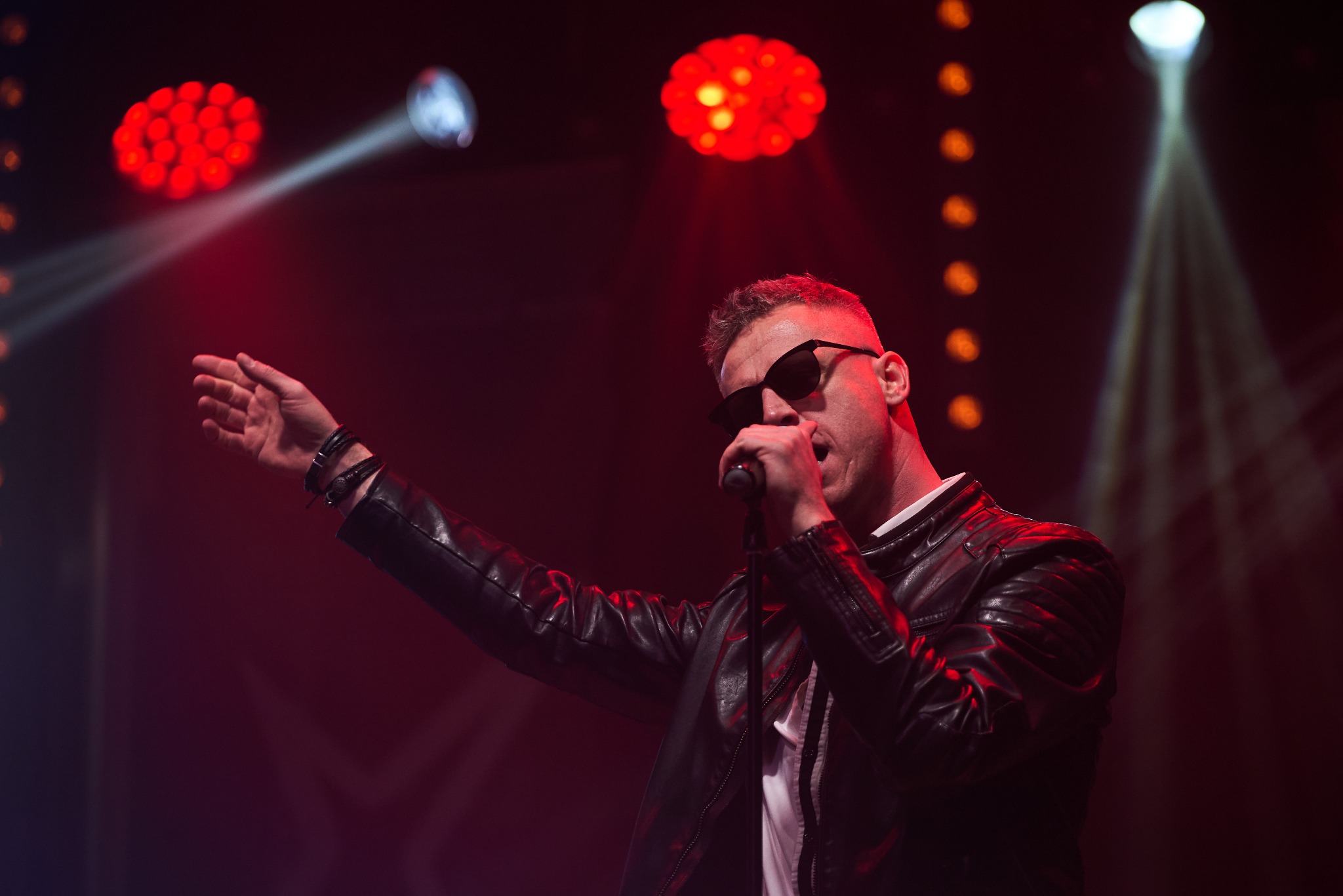 Wokalista w czarnej skórzanej kurtce i okularach przeciwsłonecznych śpiewa do mikrofonu na scenie oświetlonej czerwonymi i pomarańczowymi światłami.