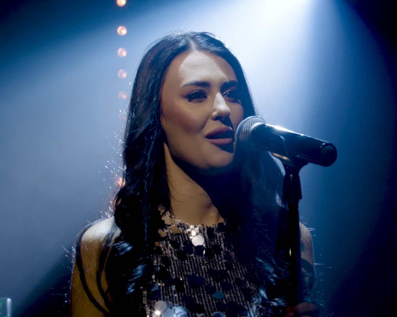 Wokalistka z długimi ciemnymi włosami śpiewa do mikrofonu na scenie w niebieskim oświetleniu punktowym, ubrana w błyszczącą sukienkę.