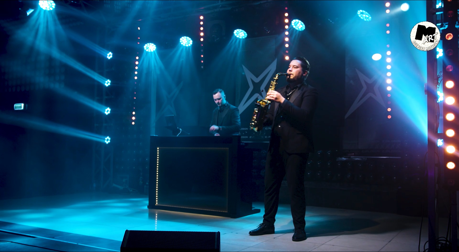 Wokalista grający na saksofonie na scenie z DJ-em w tle, oświetlenie sceniczne w kolorze niebieskim i białym, logo zespołu w prawym górnym rogu.