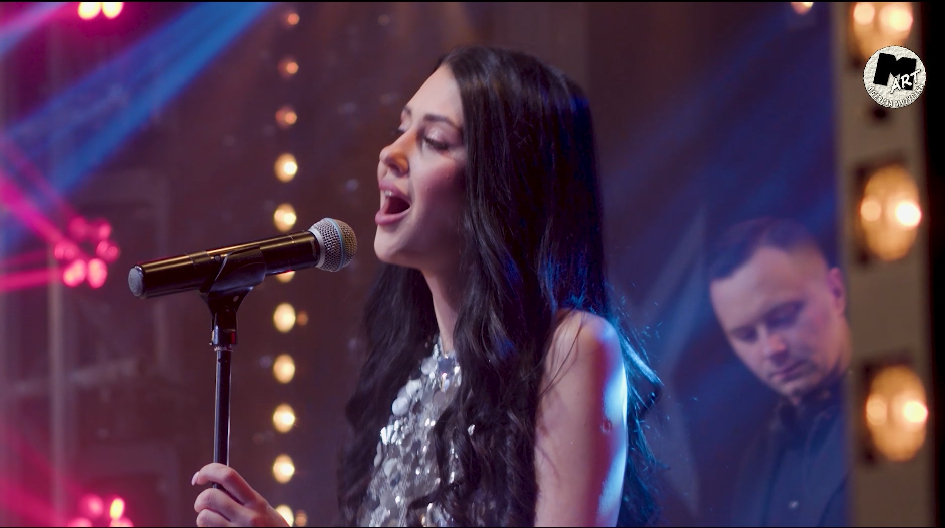 Wokalistka w srebrnej sukience śpiewa do mikrofonu na scenie z oświetleniem punktowym, w tle widoczny DJ w słuchawkach i logo agencji muzycznej.