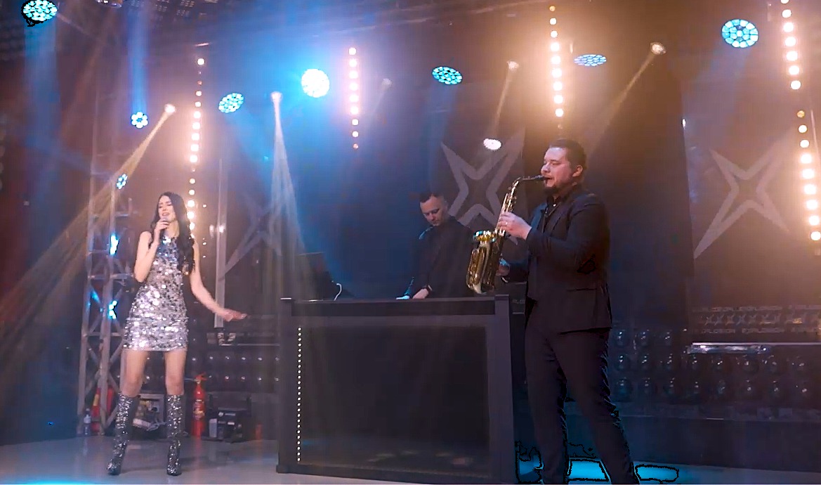 Wokalistka w srebrnej sukience i kozakach, saksofonista w garniturze i DJ za konsoletą podczas występu na scenie oświetlonej reflektorami.