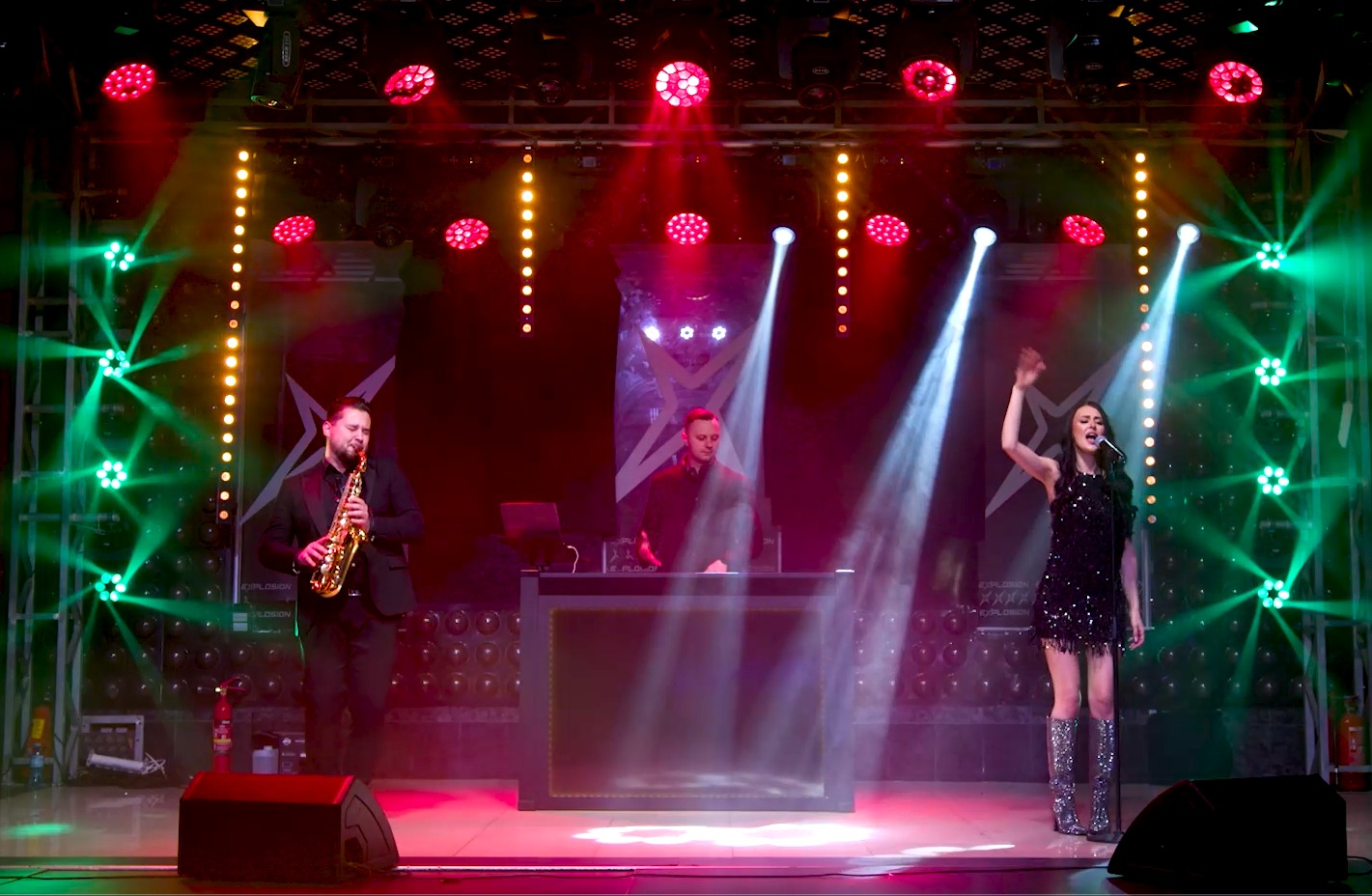 Scena z oświetleniem, na której występuje wokalistka w błyszczącej sukience i srebrnych kozakach, saksofonista w garniturze oraz DJ za konsoletą.