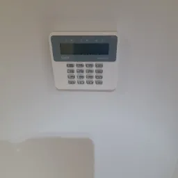 Centrala alarmowa Satel Perfecta zamontowana na białej ścianie, widok z przodu, na wyświetlaczu ciemny ekran.