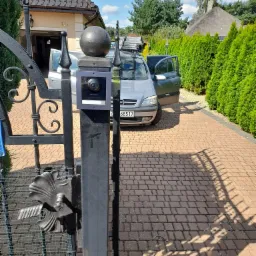Wideodomofon Hikvision zamontowany na metalowej bramie wjazdowej, z widocznym domem i zaparkowanym samochodem na podjeździe z kostki brukowej.