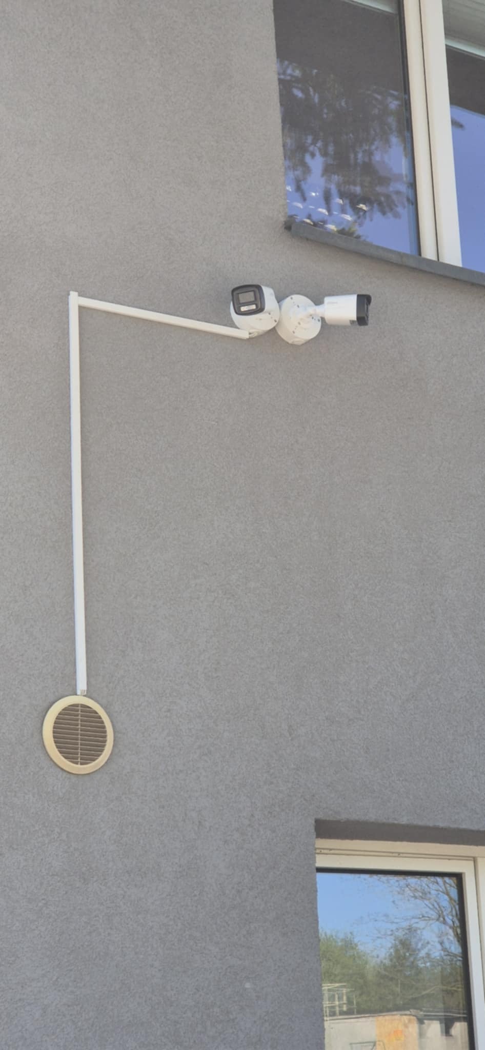 Zewnętrzny montaż dwóch kamer monitoringu na białym wysięgniku przy szarej ścianie budynku. Widoczny otwór wentylacyjny i okna z odbiciem.