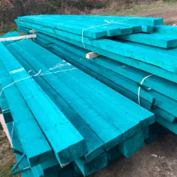 Stos turkusowej tarcicy budowlanej ułożonej na zewnątrz, zabezpieczonej białym sznurkiem, z widocznym tłem jesiennych drzew